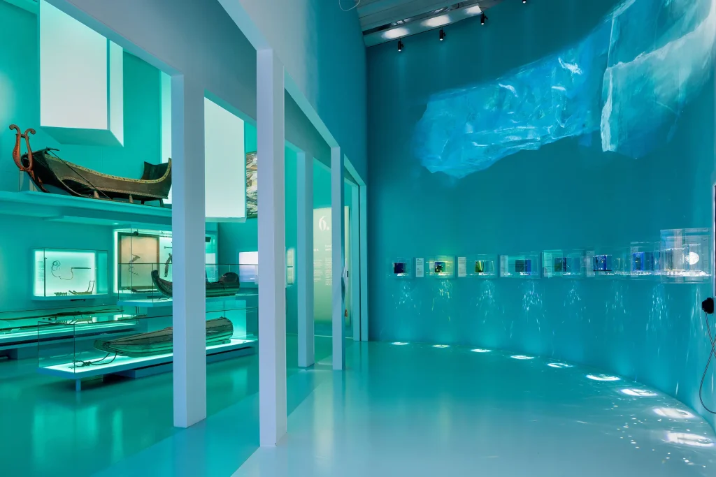 Ett turkost rum med projicerade flytblock av is på väggen, till vänster ett antal äldre kajaker, en släde och fiskeredskap.