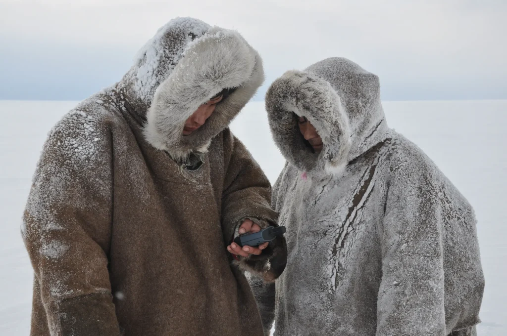 Två män står bland vita vidder klädda i snöiga anoraker med pälskantad kapuschong, den ena mannen håller i en äldre mobiltelefon eller mätare av något slag.