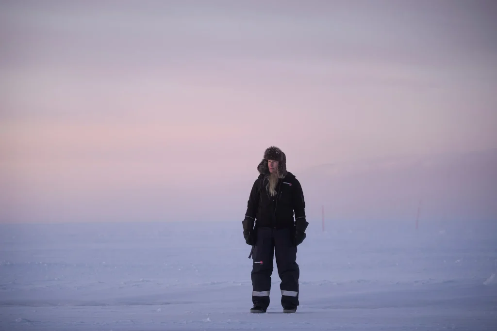 En kvinna i scooteroverall och pälsmössa står ensam på isen som är täckt av snö.