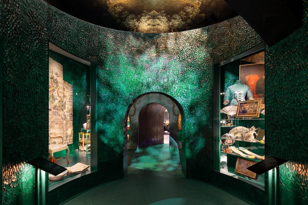 Ett omslutande rum vars tapet och väggar föreställer grönska och växtlighet, i mitten en portal och slingrande gång som leder förbi montrar med föremål från 1700-talet.