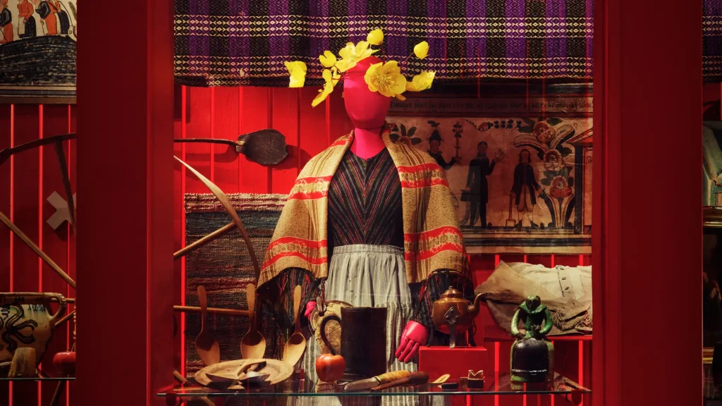 En skyltdocka med en pigas klänning i halvylle och sjal, framför dockan syns många vardagsföremål ur vardagslivet, bonader och mönstrade vävda textilier, allt har starka färger i rött, gult och lila.