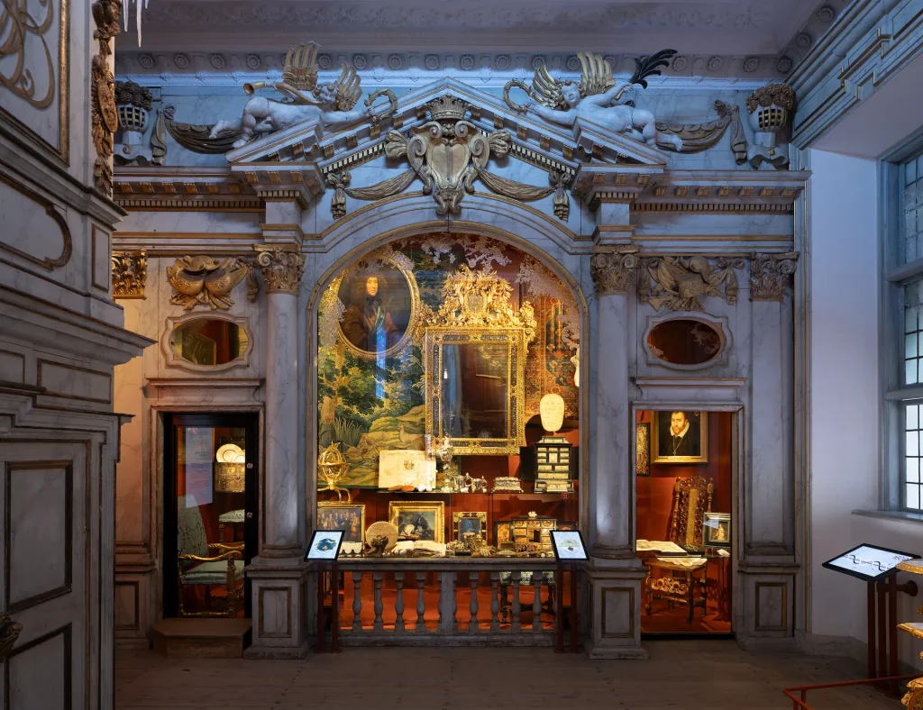 Ett stort rum 1600-talsstil i kallt ljus, i en monter syns påkostade målningar, smycken, möbler och andra föremål.