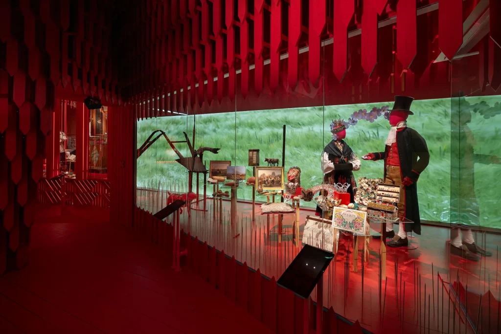 Ett starkt rött rum med en scenografi med träribbor, en lång monter med textilföremål och slöjdhantverk, två dockor i färgstarka folkdräkter, i fonden ett böljande sädesfält.