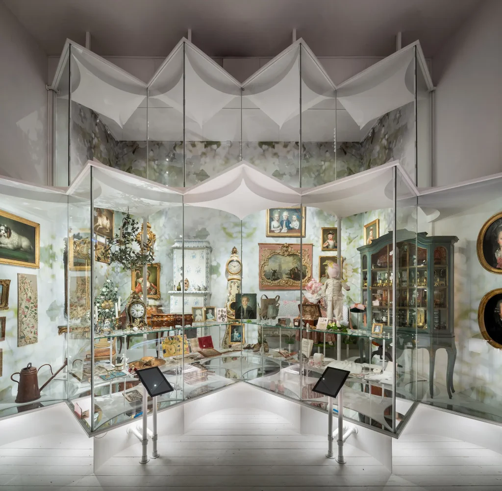 Ett ljust utställningsrum med massor av föremål från 1700-talet. I fonden en tapet som föreställer äppelblom i närbild.