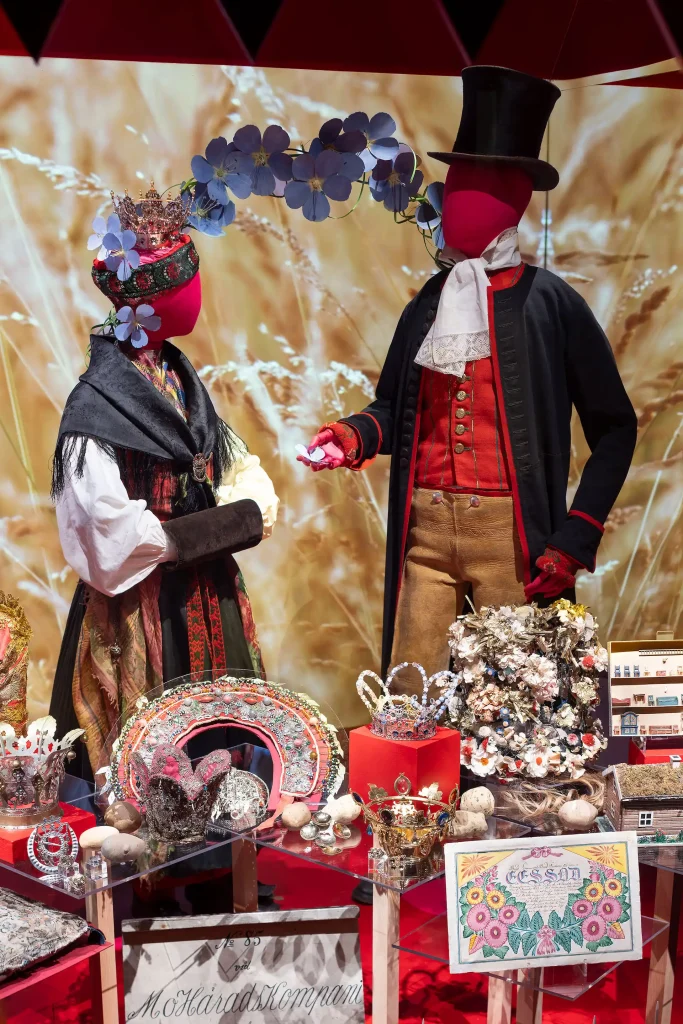 En monter med två dockor klädda i högtidskläder från 1800-talets bondesamhälle. Kläderna har starka färger och framför dockorna finns många hemslöjdade föremål som brudkronor och andra smycken.