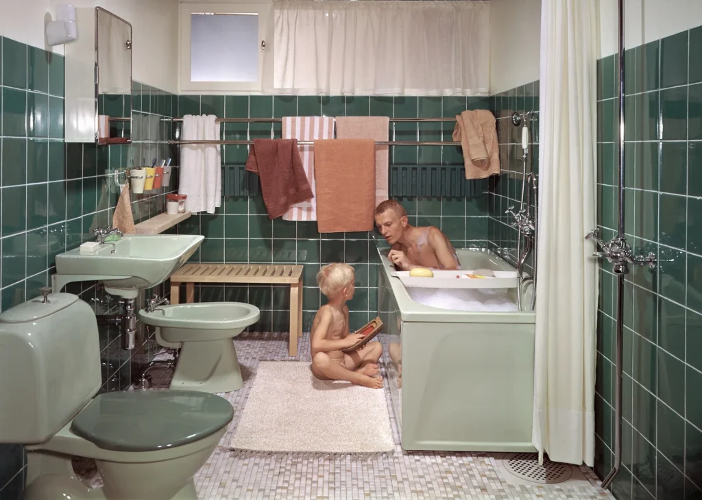 Ett grönkaklat badrum med fönster, badrumsspegelskåp, porslinslampett där både bidé, tvättställ, wc-stol och badkar går i pastellgrönt. En man badar skumbad i karet och på badrumsmattan sitter en liten pojke med och leker med en båt.