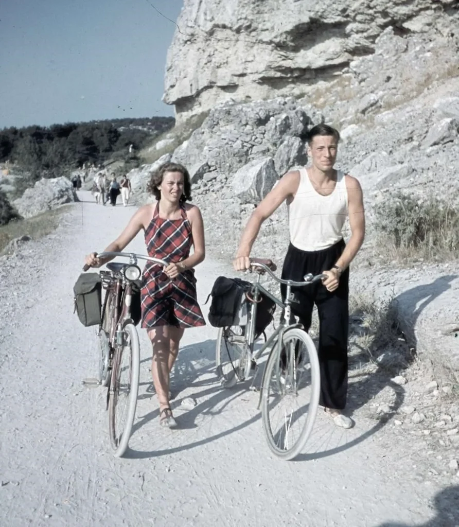 En ung kvinna i rutig solklänning och en man i vitt linne och långbyxor leder sina cyklar på en kalkstensdammig grusväg längs gotländska klintväggar.