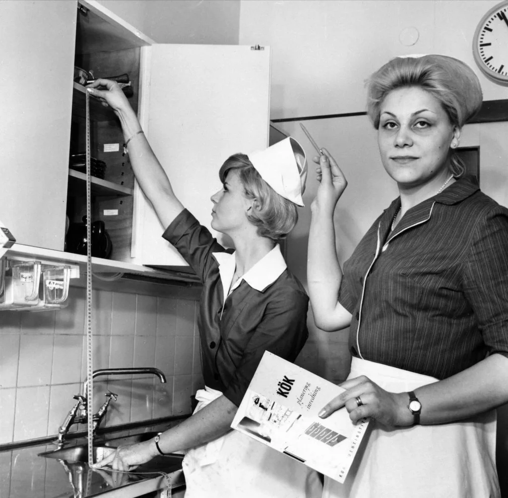 Två kvinnor med vita förkläden och hättor mäter avstånd mellan diskho och köksskåp i ett funktionalistiskt kök