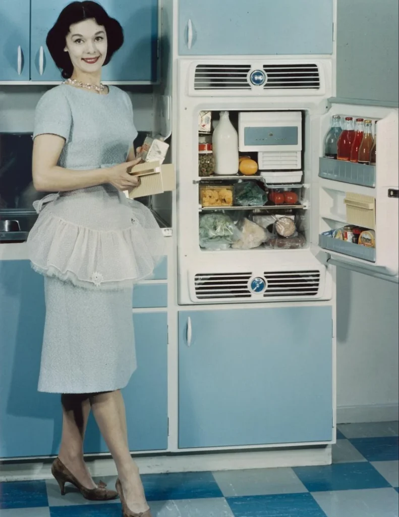 En kvinna poserar i ett pastellblått kök vid ett fyllt kylskåp med frysfack, hon är klädd i klänning, förkläde och pumps och bär halsband och örhängen till rött läppstift.