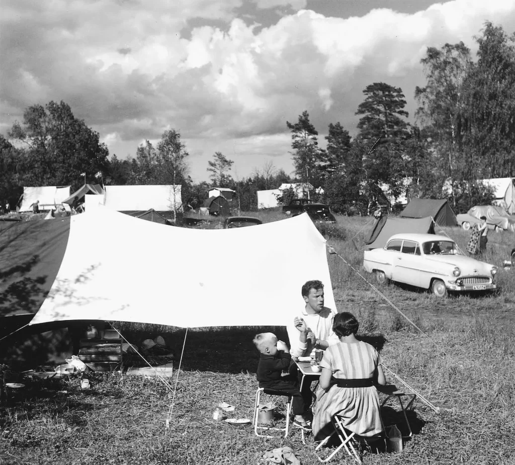 En campingplats på 1950- eller 1960-talet med tält, en volvo Amazon och en familj sitter framför sitt tält vid campingbord, kvinnan i randig bomullsklänning, mannen röker en cigarett och en liten pojke dricker ur en mugg.