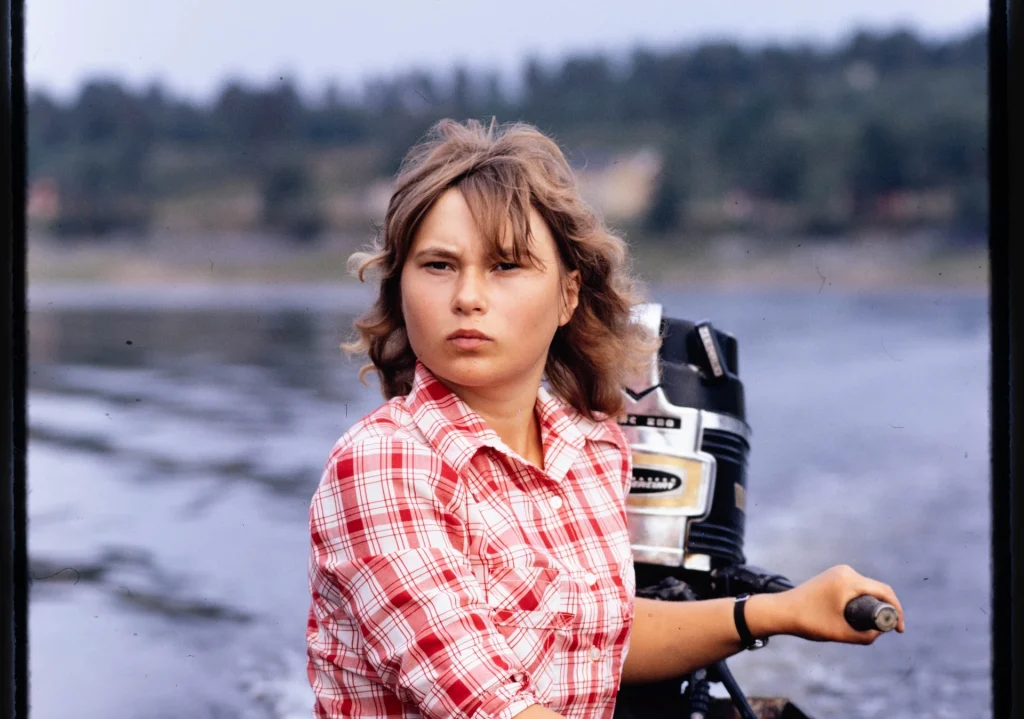 En flicka i vitrutig skjorta sitter vid rodret och styr en motorbåt, hon ser koncentrerad ut.