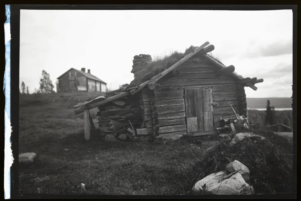 En svartvit bild föreställande en knuttimrad bod på en höjd vid vattnet.