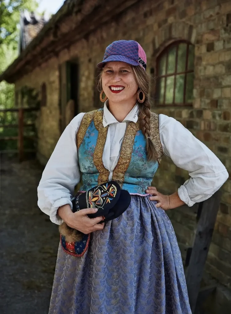 En kvinna i folkdräkt i blå nyanser med keps, fläta, ringar i öronen och röda läppar håller broderade vantar i handen, i bakgrunden en tegelbyggnad.